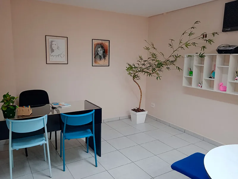 Consultório da clínica, com mesa e duas cadeiras azuis, plantas e quadros na parede