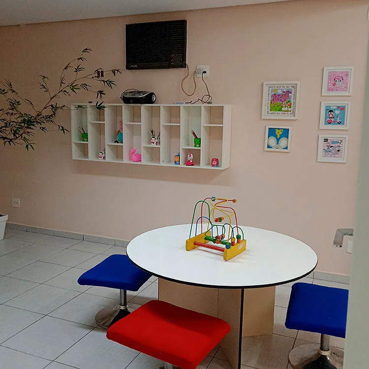 Sala de atendimento da clínica, com mesa redonda baixa, banquetas coloridas e estante com brinquedos