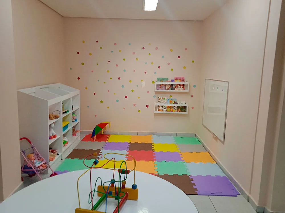 Sala lúdica da Clínica Thetis, com tapete de peças coloridas, estante de brinquedos e mesa redonda de atividades